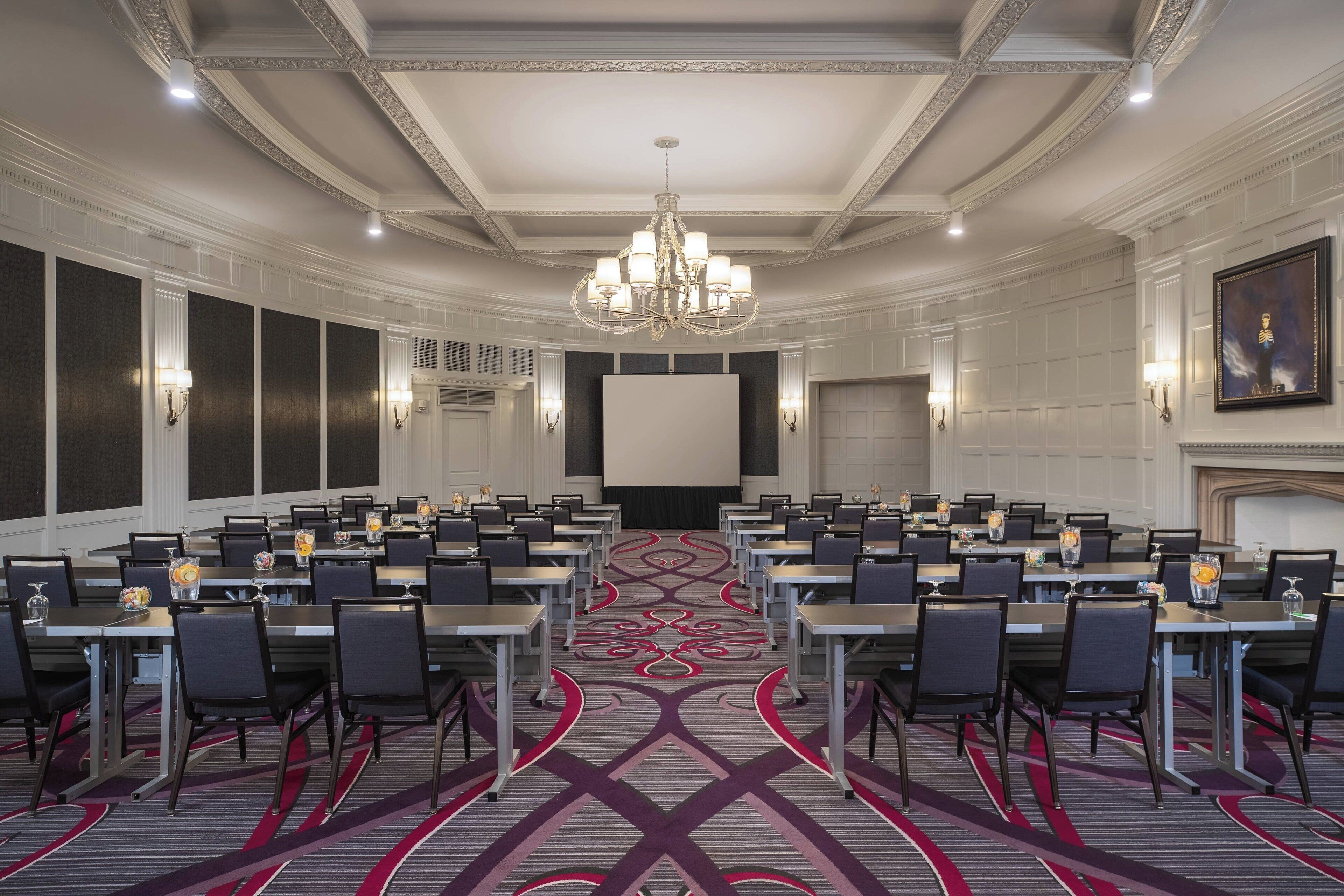A conference room with a large screen, multiple tables, and chairs, featuring a patterned carpet and a chandelier.