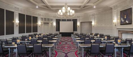 A conference room with a large screen, multiple tables, and chairs, featuring a patterned carpet and a chandelier.