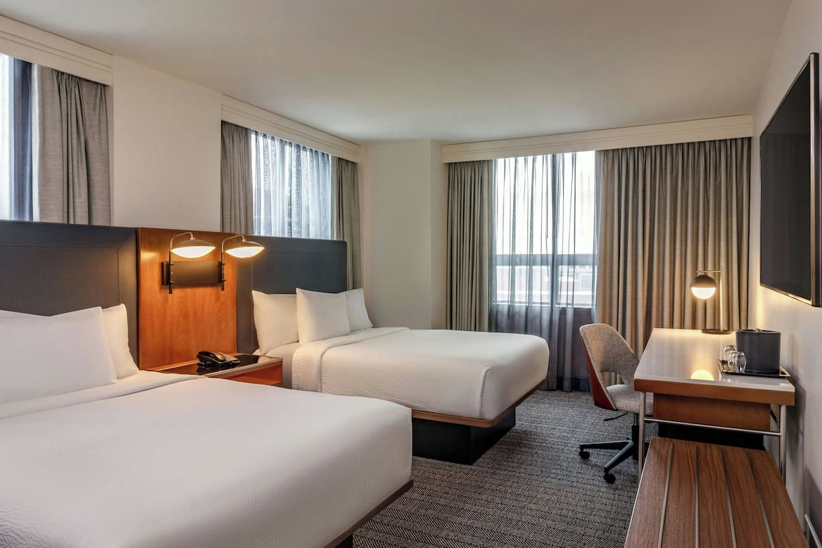 Quarto de hotel com duas camas, uma escrivaninha, uma cadeira, uma televisão e um telefone na mesinha de cabeceira.