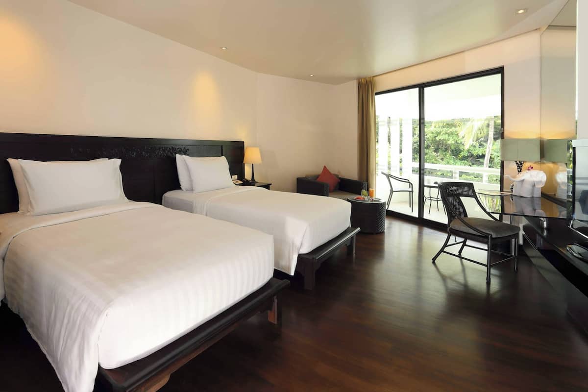A hotel room with two beds, a small table, a chair, and a balcony with a view.