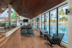 A spacious indoor area with a wooden ceiling, large windows overlooking a pool and palm trees, and modern seating arrangements.