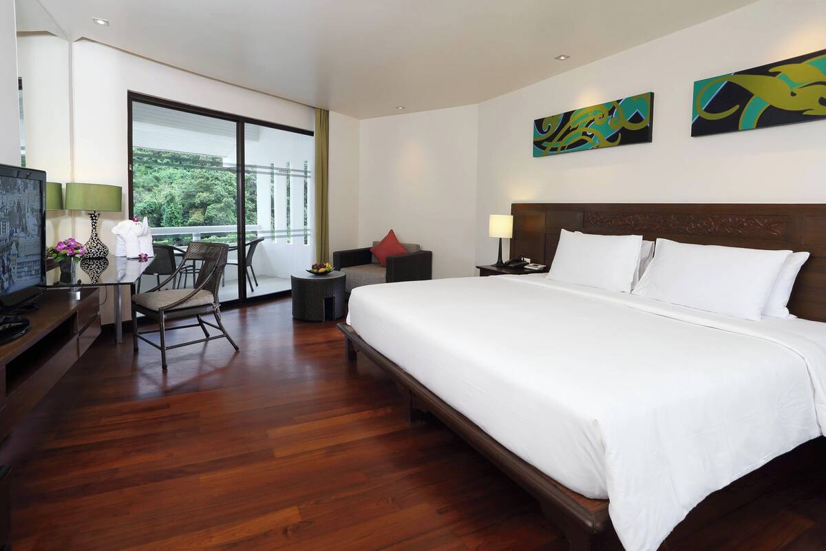 A hotel room with a large bed, a desk with a computer, a chair, a small table with a lamp, and a view of greenery through the window.
