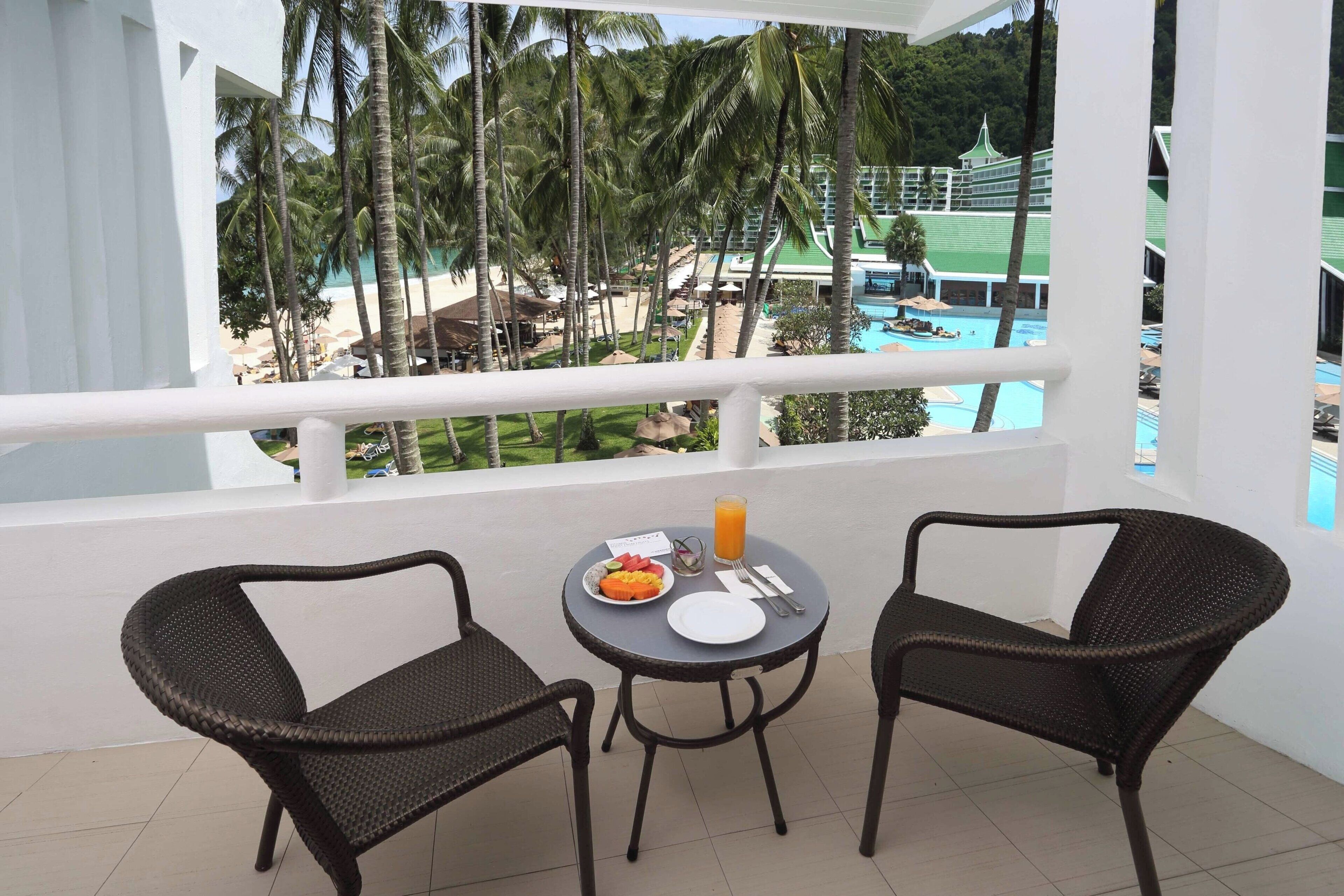 A balcony with two wicker chairs, a small round table with a plate, a glass of orange juice, and a menu. The view includes a pool, palm trees, and a building with a green roof.