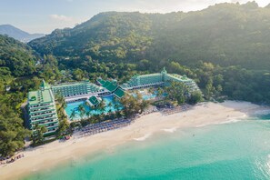 Vista aérea de un resort con varias edificaciones de techo verde, una playa y una piscina.