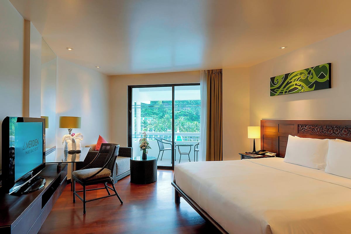 A hotel room with a bed, a TV, a chair, a small table with a lamp, and a balcony with a view of greenery.