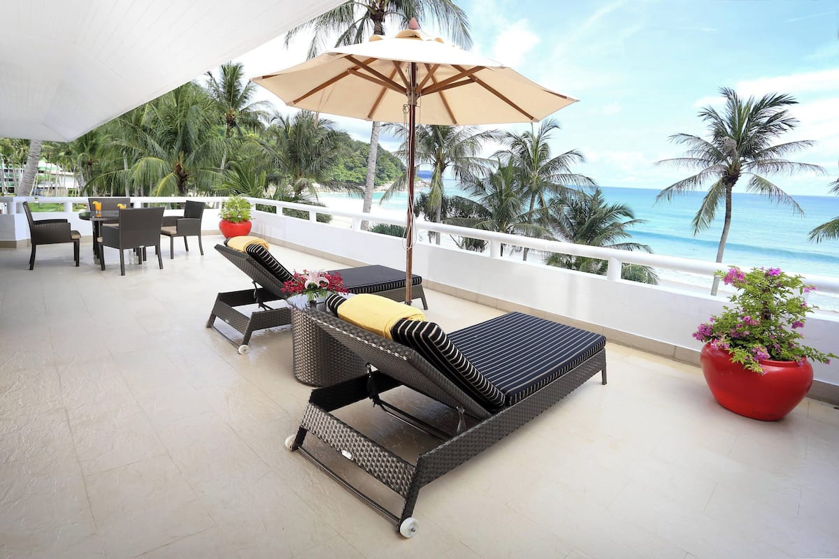 A sun lounge with a table and chairs, a large umbrella, and a view of the ocean and palm trees.