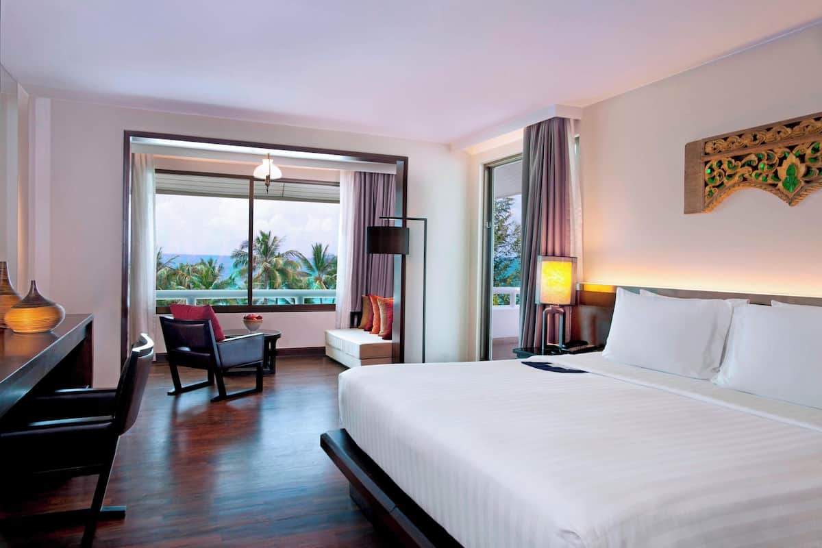 A hotel room with a bed, a desk, a chair, a window with a view of palm trees, and a nightstand with a lamp.