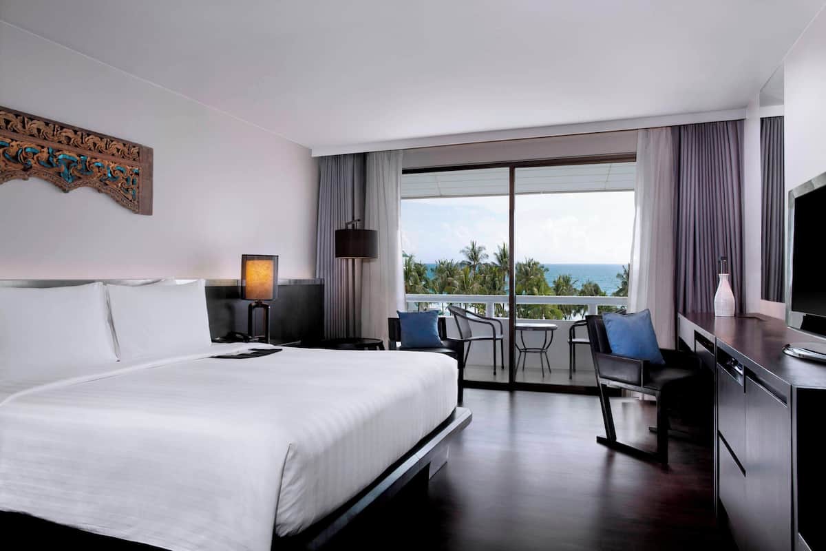 A hotel room with a large bed, a desk with a chair, a TV, and a balcony with a view of the ocean.