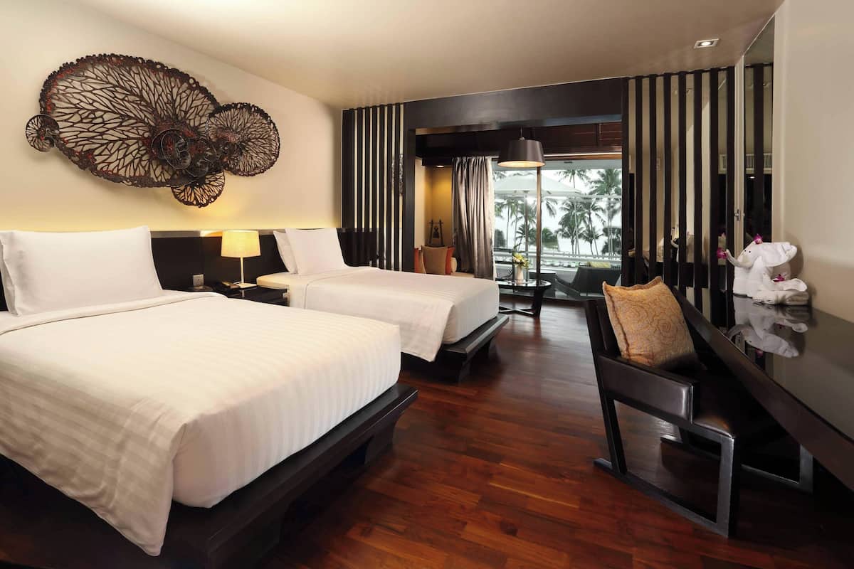 A hotel room with two beds, a large decorative wall piece, a dining area with a table and chairs, and a view of palm trees through the window.