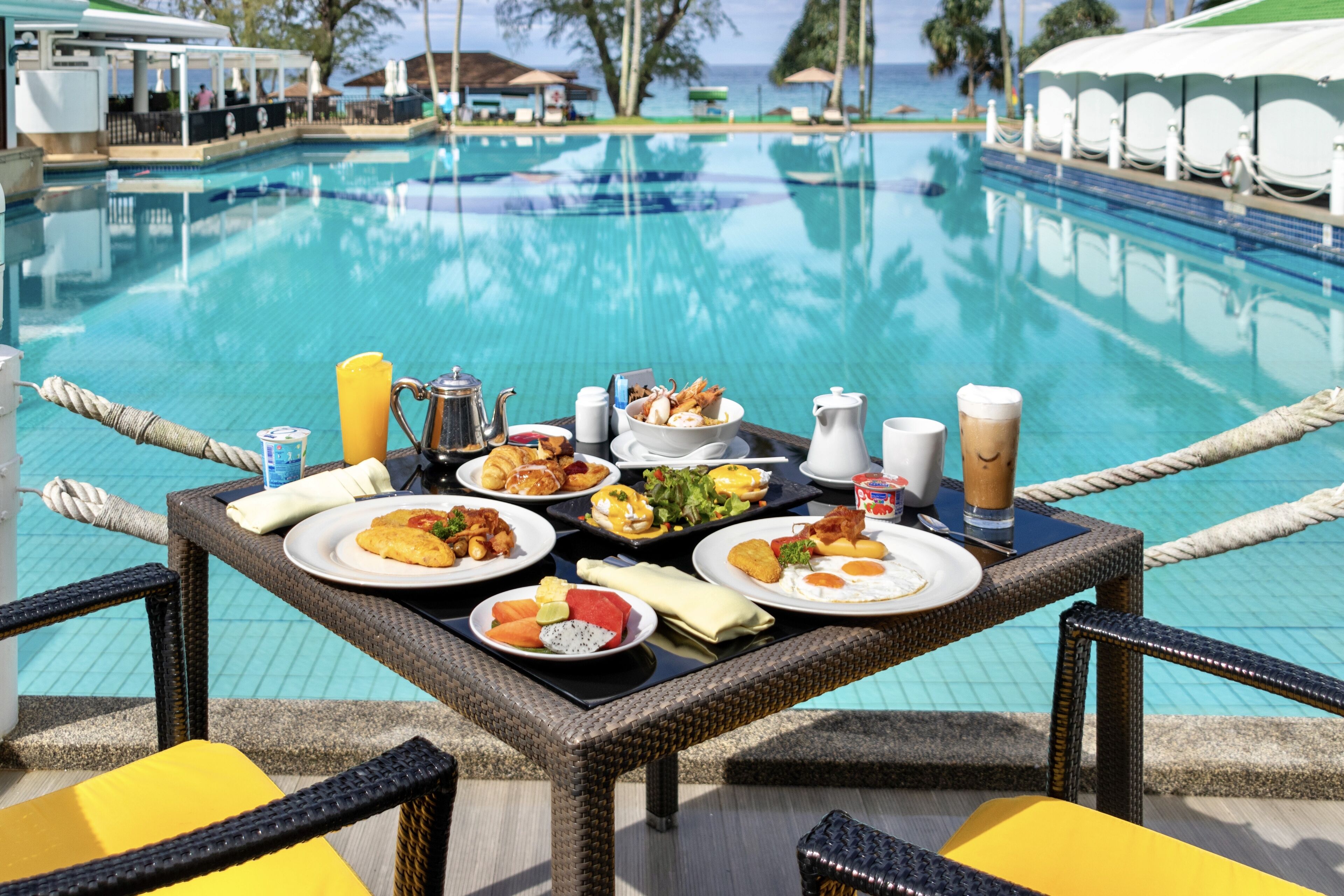 A dining table with various dishes and drinks by a poolside, overlooking the water.