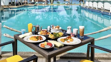 A dining table with various dishes and drinks by a poolside, overlooking the water.