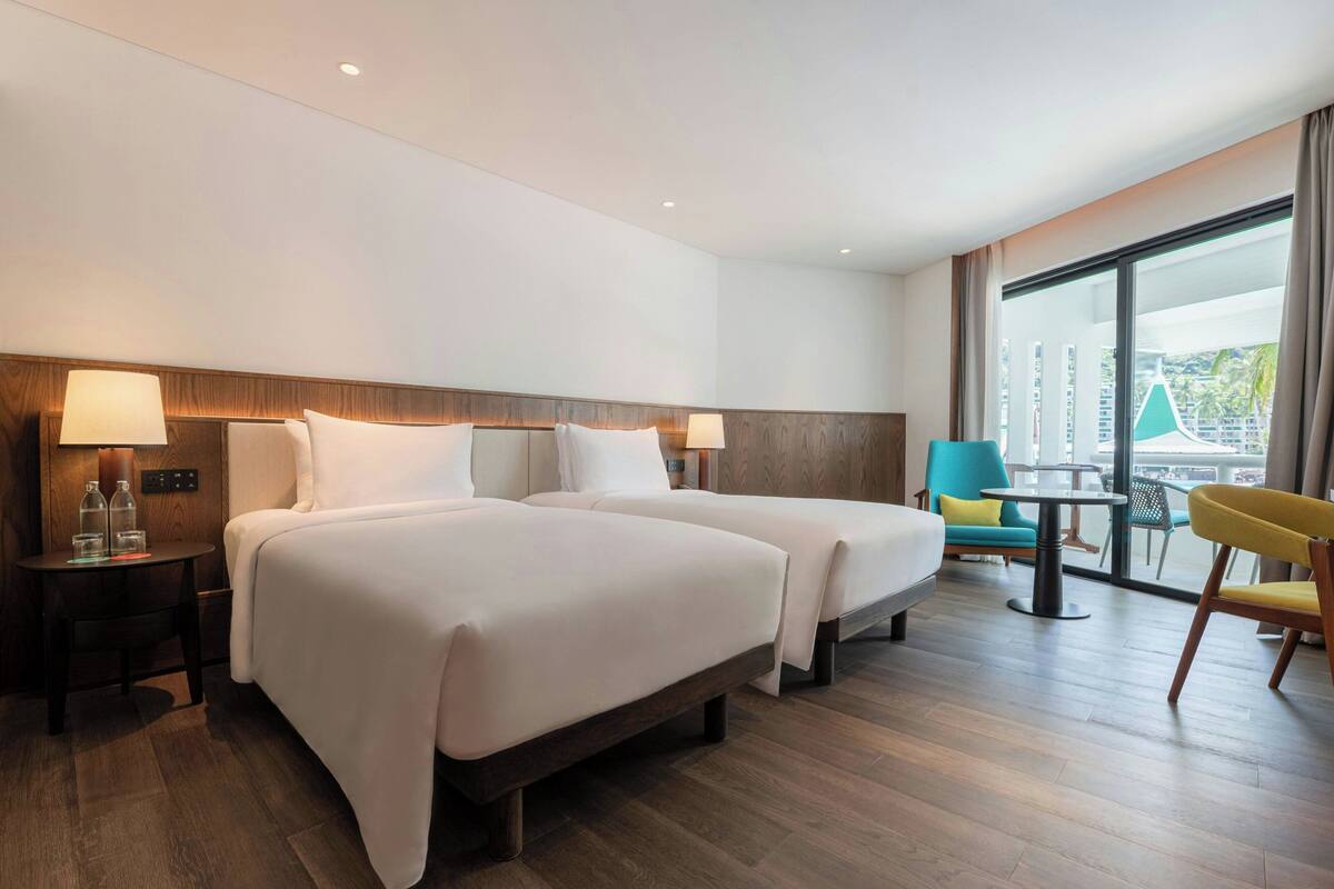 A hotel room with two beds, a small table, a chair, and a view of the city.