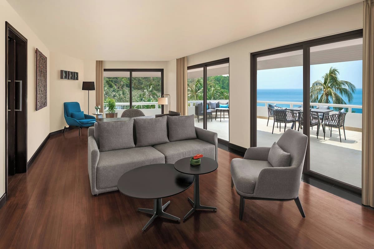 A modern hotel room with a grey sofa, two armchairs, a small round table, and a balcony with a view of the ocean.