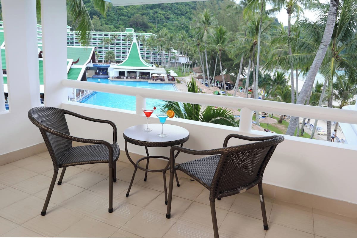 A balcony with two wicker chairs and a small round table, overlooking a pool and a resort area with palm trees and a building with a green roof.