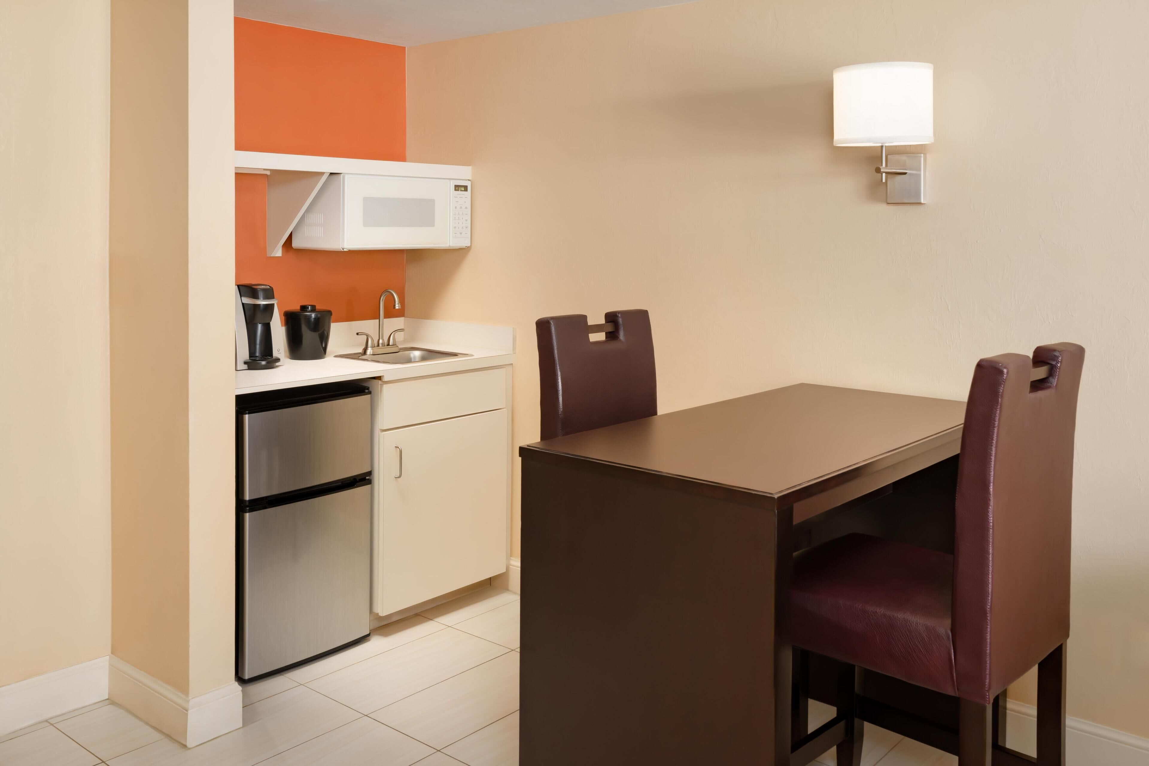 Une cuisine compacte équipée d’un four à micro-ondes, d’une machine à café et d’un réfrigérateur, ainsi qu’une table à manger avec deux chaises et un luminaire fixé au mur.