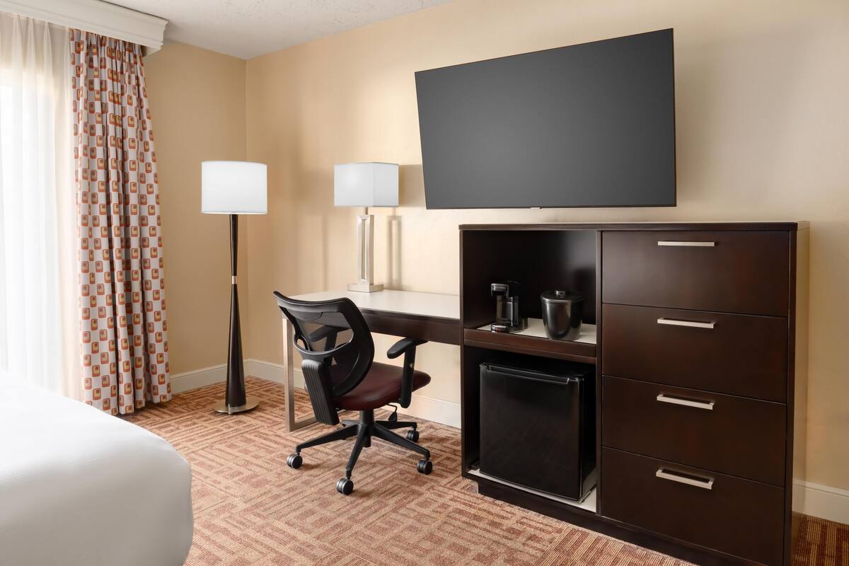 A hotel room with a bed, a desk with a chair, a flat-screen TV, a lamp, and a minibar.