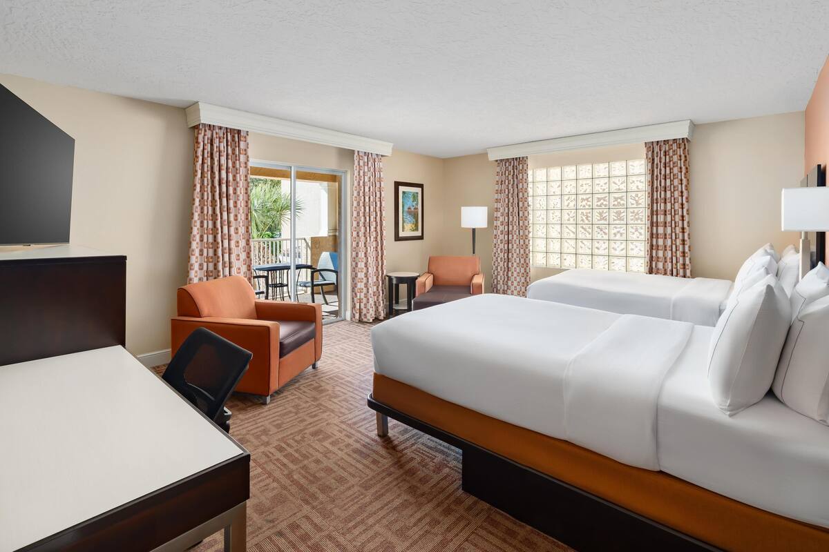 A hotel room with a bed, a desk, an orange chair, and a view of the outdoors.