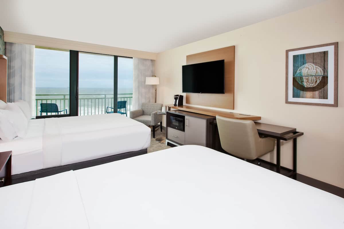 A hotel room with two beds, a desk, a chair, a TV, and a large window offering a view of the ocean.