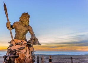 A large bronze statue of a muscular figure holding a trident, situated on a rocky pedestal by the beach.