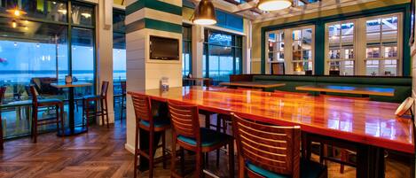 A bar area with wooden chairs and a long bar counter, large windows with a view, and a ceiling with wooden beams.