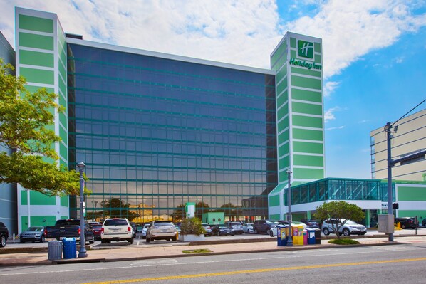 A Holiday Inn hotel with a modern glass facade, a parking lot, and a pedestrian walkway.