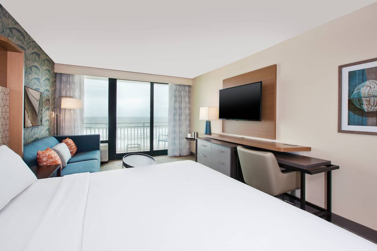 A hotel room with a large bed, a desk, a chair, a television, and a view of the ocean through the window.