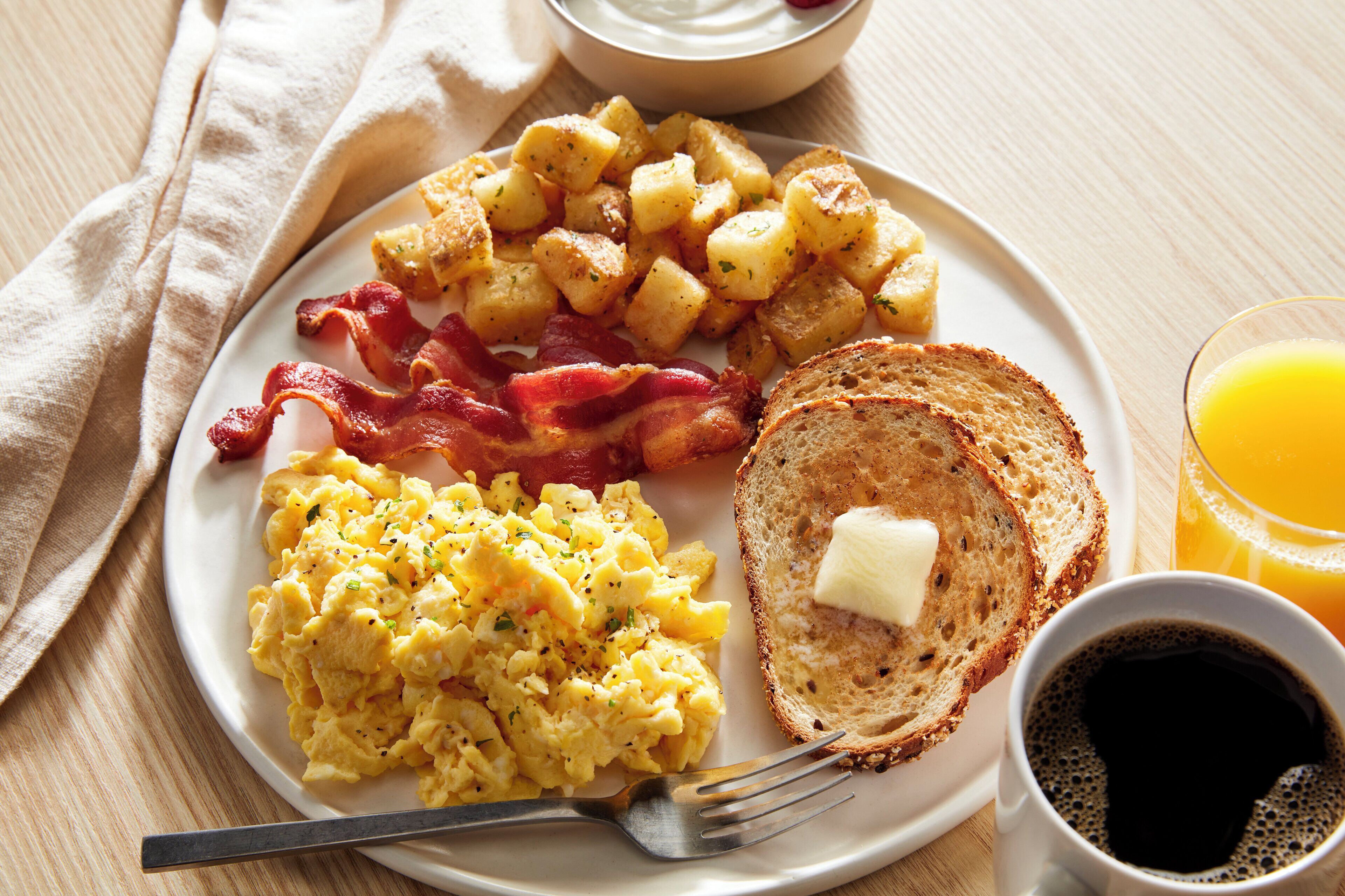 A plate with scrambled eggs, bacon, roasted potatoes, toast with butter, and a glass of orange juice.