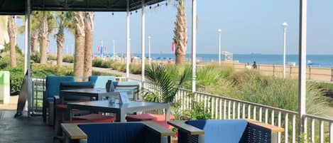 A covered outdoor seating area with tables and chairs, overlooking a beach with palm trees and a clear sky.