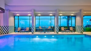 Piscina interna com água azul cristalina, rodeada por espreguiçadeiras e mesas, com amplas janelas oferecendo vista do horizonte urbano.