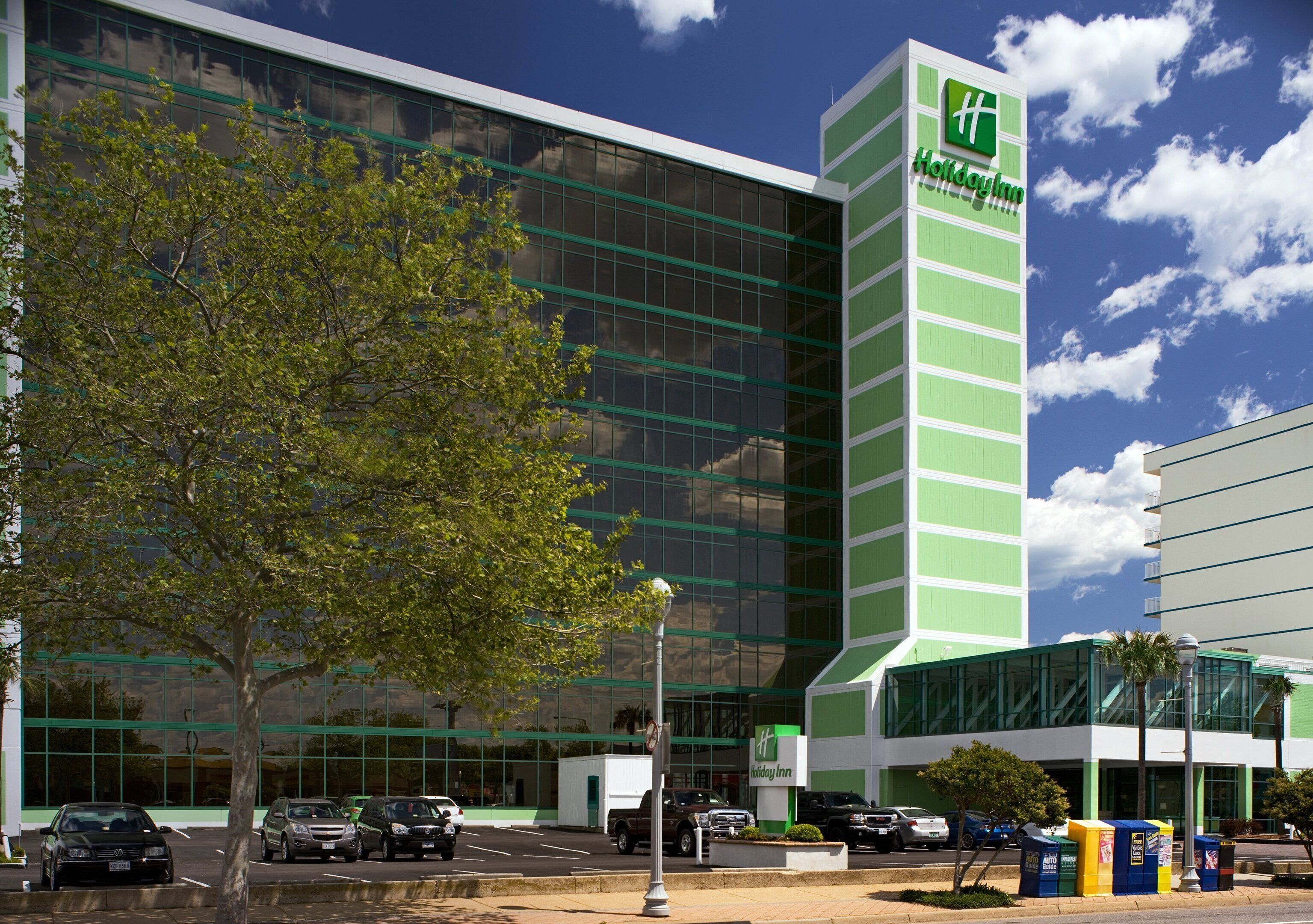 Um hotel Holiday Inn com fachada de vidro moderna, um esquema de cores marcante em verde e branco, e um estacionamento com vários carros.