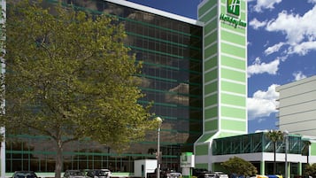 Um hotel Holiday Inn com fachada de vidro moderna, um esquema de cores marcante em verde e branco, e um estacionamento com vários carros.