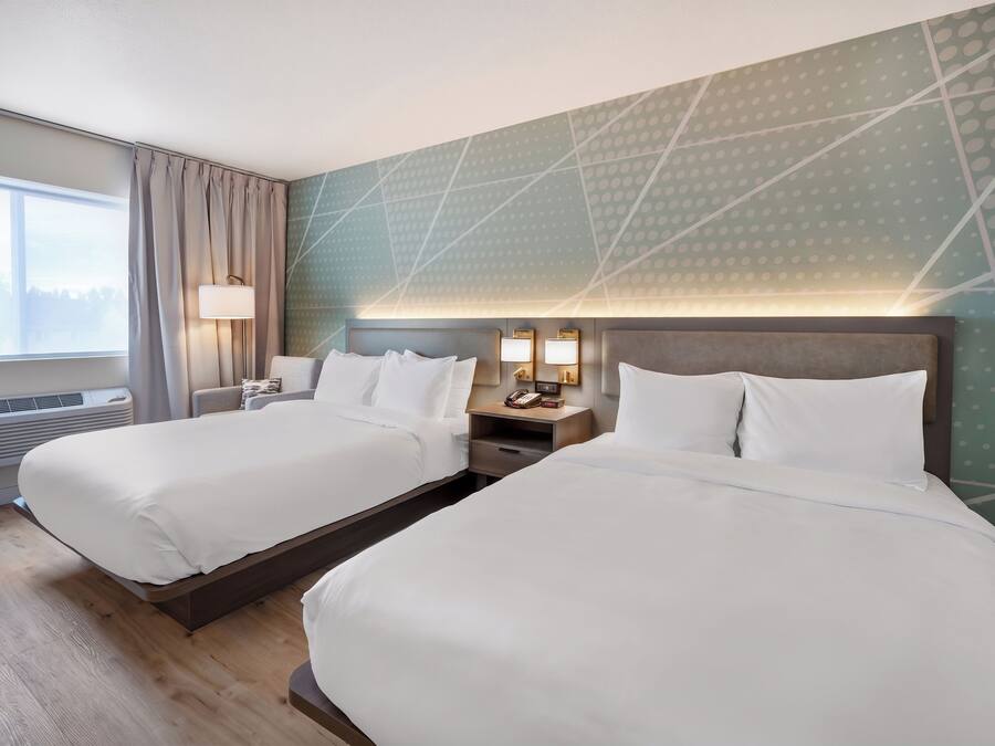 A hotel room with two beds, a nightstand with lamps, a window with curtains, and a patterned wall.