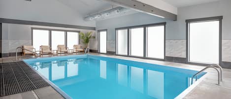 A large indoor swimming pool with clear blue water, surrounded by windows and a seating area with chairs.