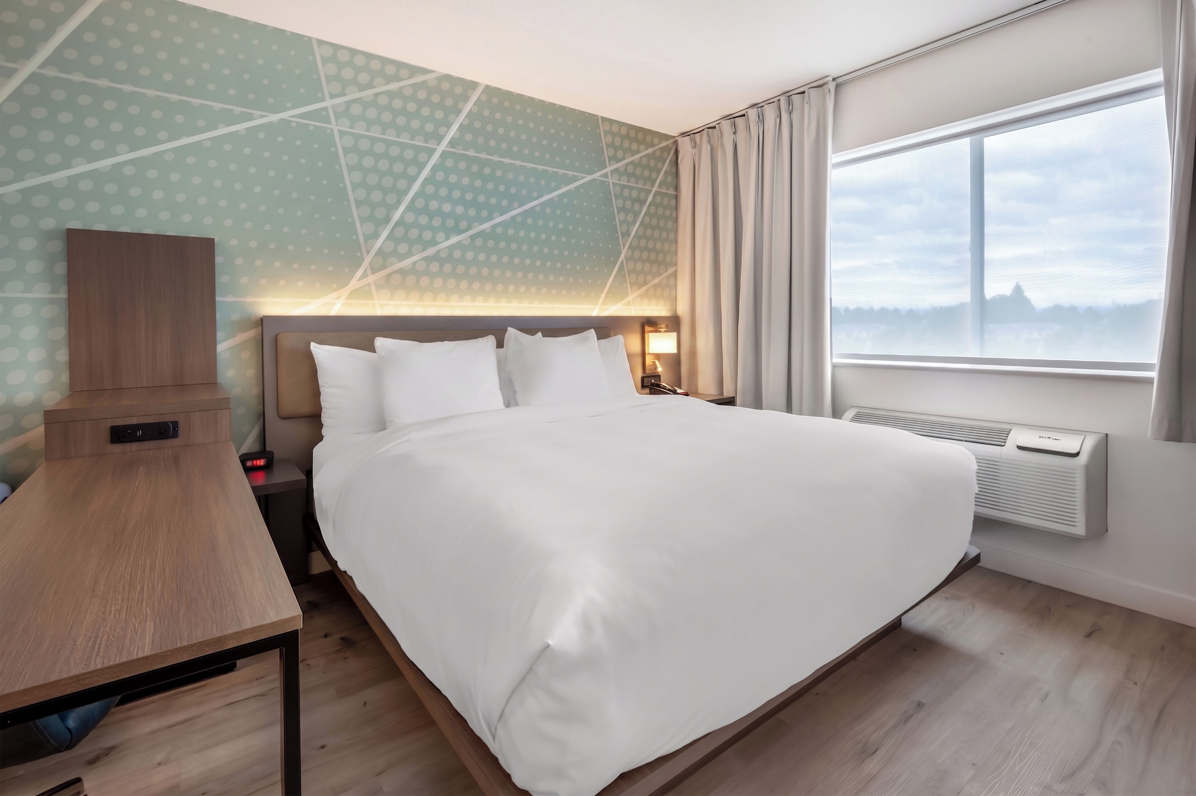 A hotel room with a bed, bedside table, lamp, and a view of the outdoors through a window.