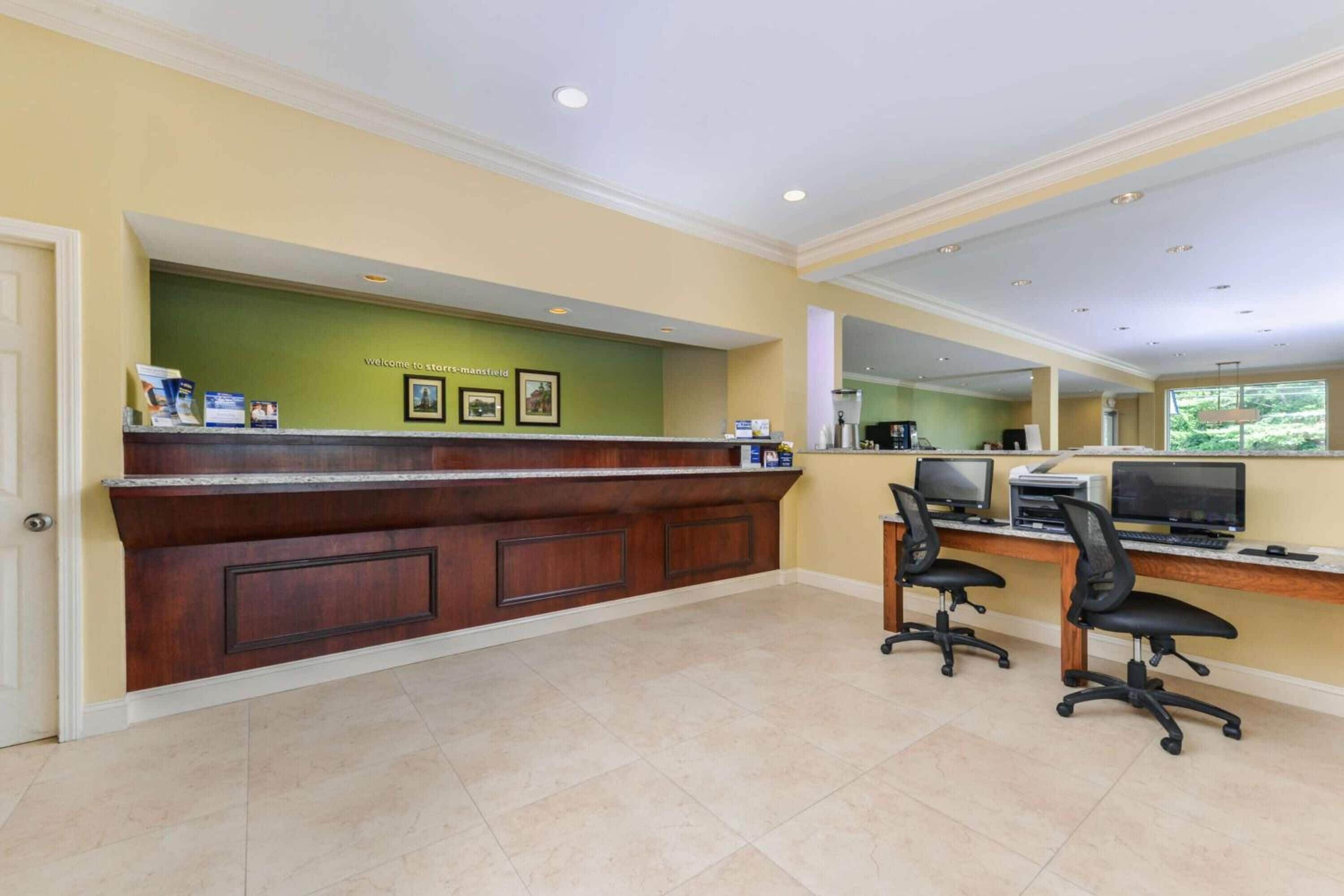 A reception area with a wooden counter, two office chairs, and a computer desk with monitors and printers.