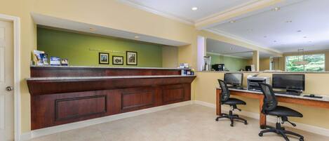 A reception area with a wooden counter, two office chairs, and a computer desk with monitors and printers.