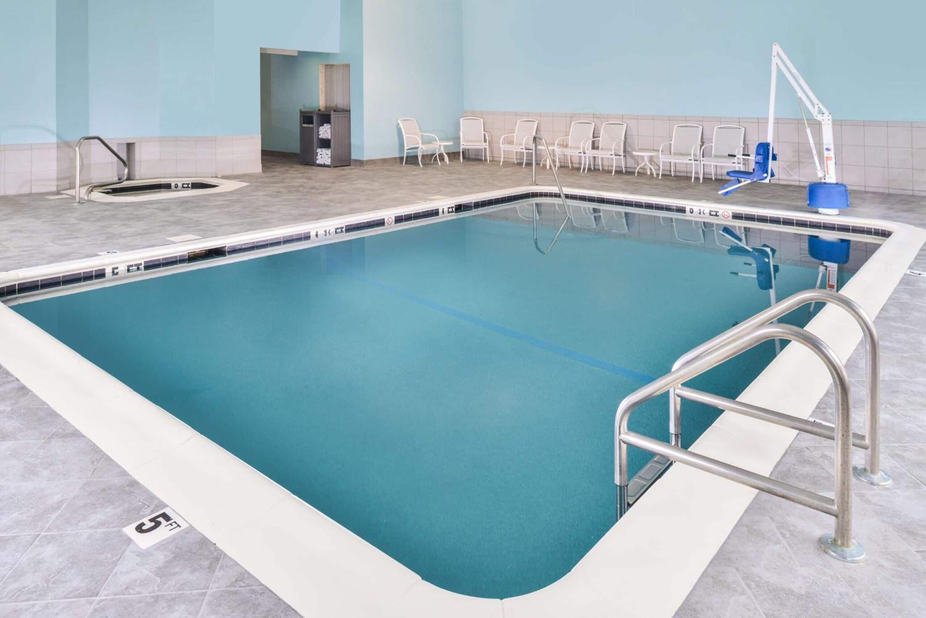Una piscina rectangular interior con superficie de agua azul cristalina, una escalera de metal y suelo de baldosas con indicador de profundidad.