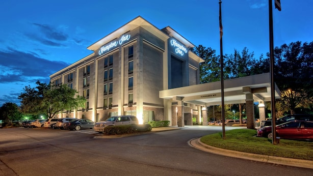A modern hotel with a prominent "Hampton Inn" sign, a covered entrance, and a parking lot with several cars.