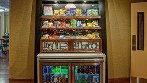 A vending machine with snacks and drinks, located in a hallway with wooden flooring and a brown wall.