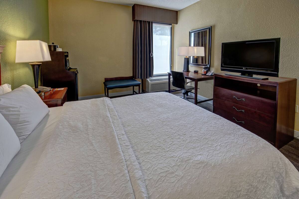 Ein Hotelzimmer mit einem großen Bett, einem Schreibtisch mit Stuhl, einem Fernseher und einem Fenster mit Vorhängen.