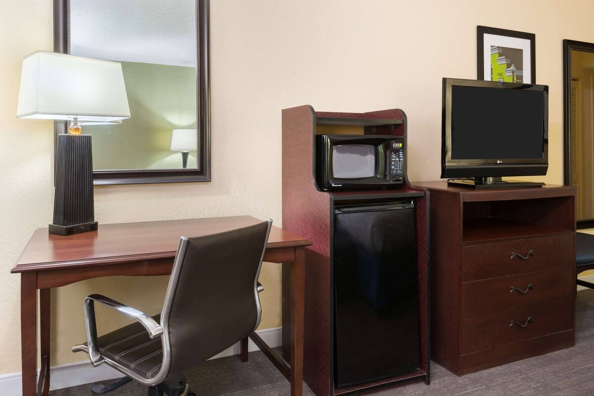 Ein Hotelzimmer mit einem Holztisch, einem modernen Sessel, einem schwarzen Kühlschrank, einer Mikrowelle, einem Flachbildfernseher und einer Lampe.