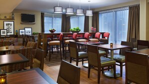 A hotel dining area with multiple tables and chairs, a television mounted on the wall, and large windows with curtains.