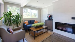 Un salon moderne avec un canapé, une table basse et une cheminée. On y trouve des plantes en pot et une vue sur l’extérieur.