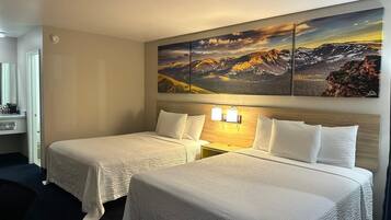 A hotel room with two beds, a large mountain landscape painting above the beds, and a nightstand with two wall-mounted lamps.