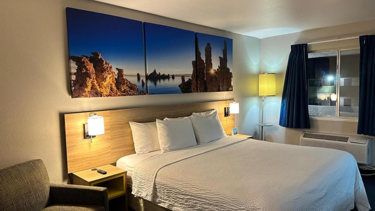 A hotel room with a large bed, two bedside lamps, a nightstand, a chair, a window with curtains, and a wall-mounted artwork.