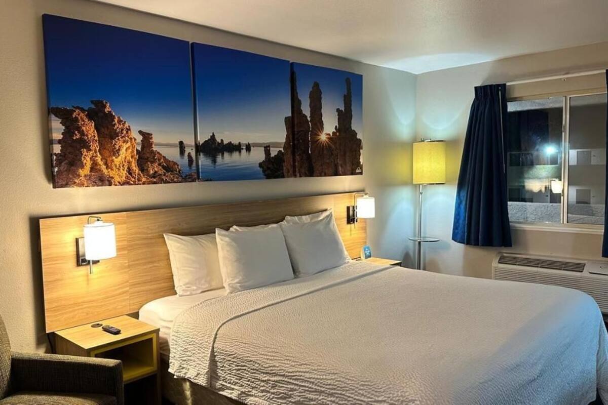 A hotel room with a large bed, two bedside lamps, a nightstand, a chair, a window with curtains, and a wall-mounted artwork.