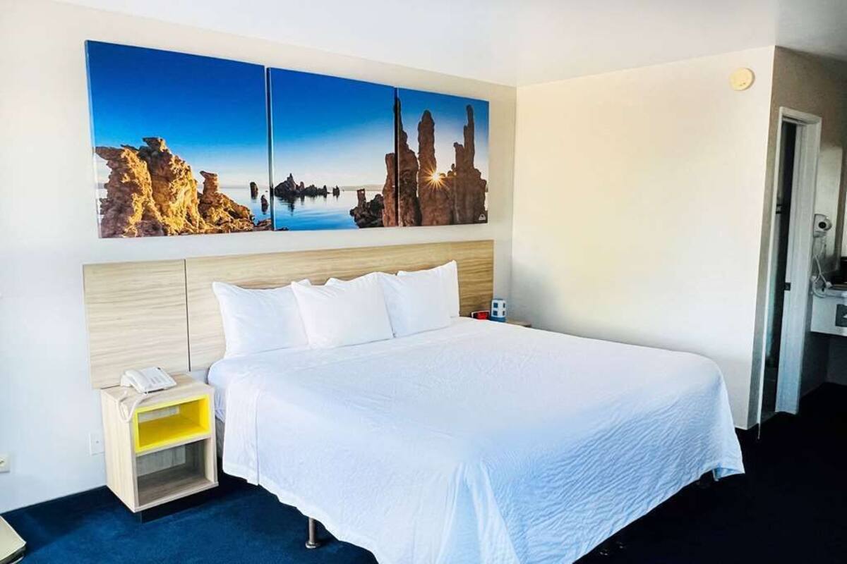 A hotel room with a large bed, a bedside table with a lamp, and a painting of a coastal landscape on the wall.
