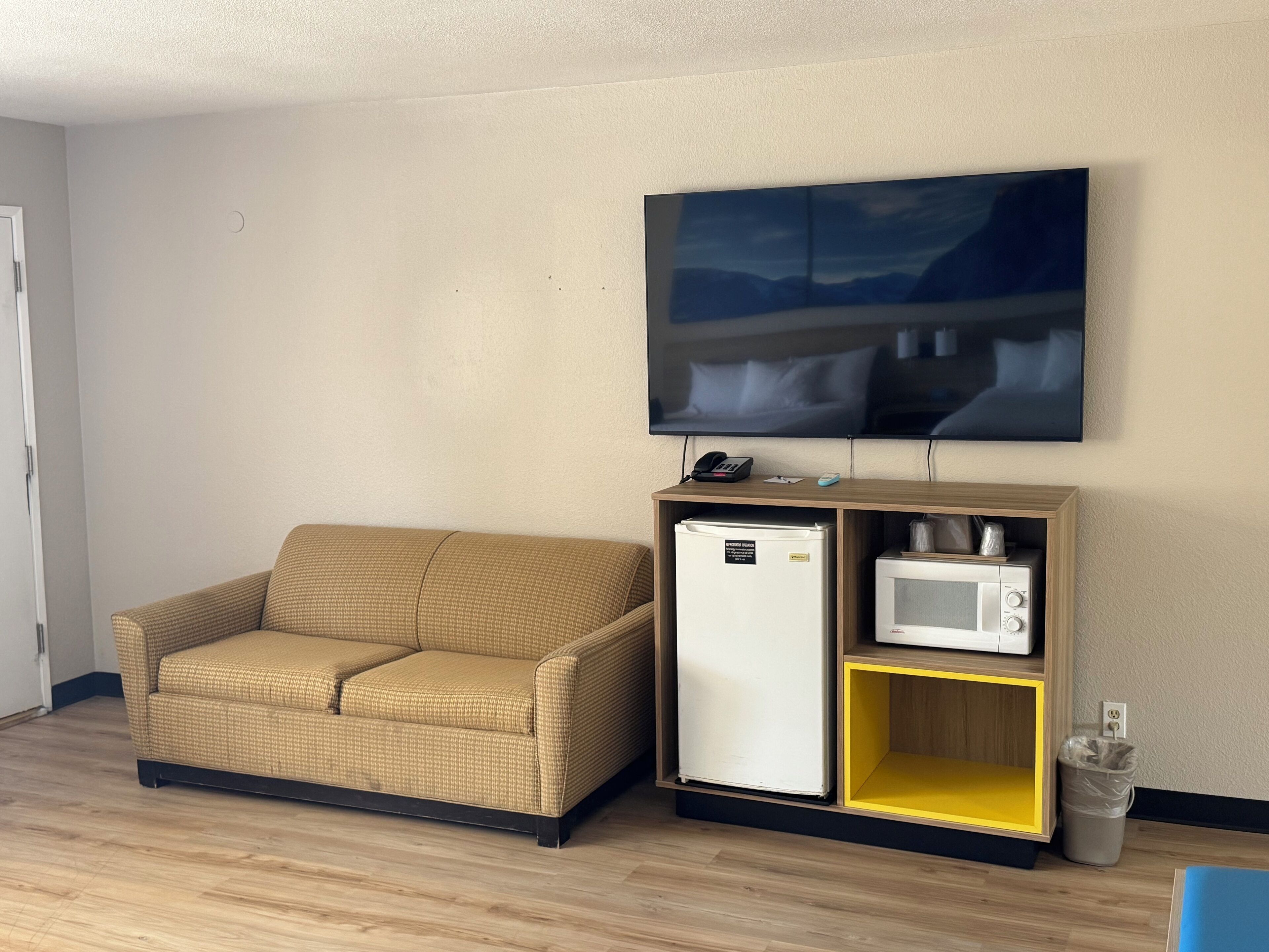 A small living area with a sofa, a mini-fridge, a microwave, and a TV mounted on the wall.