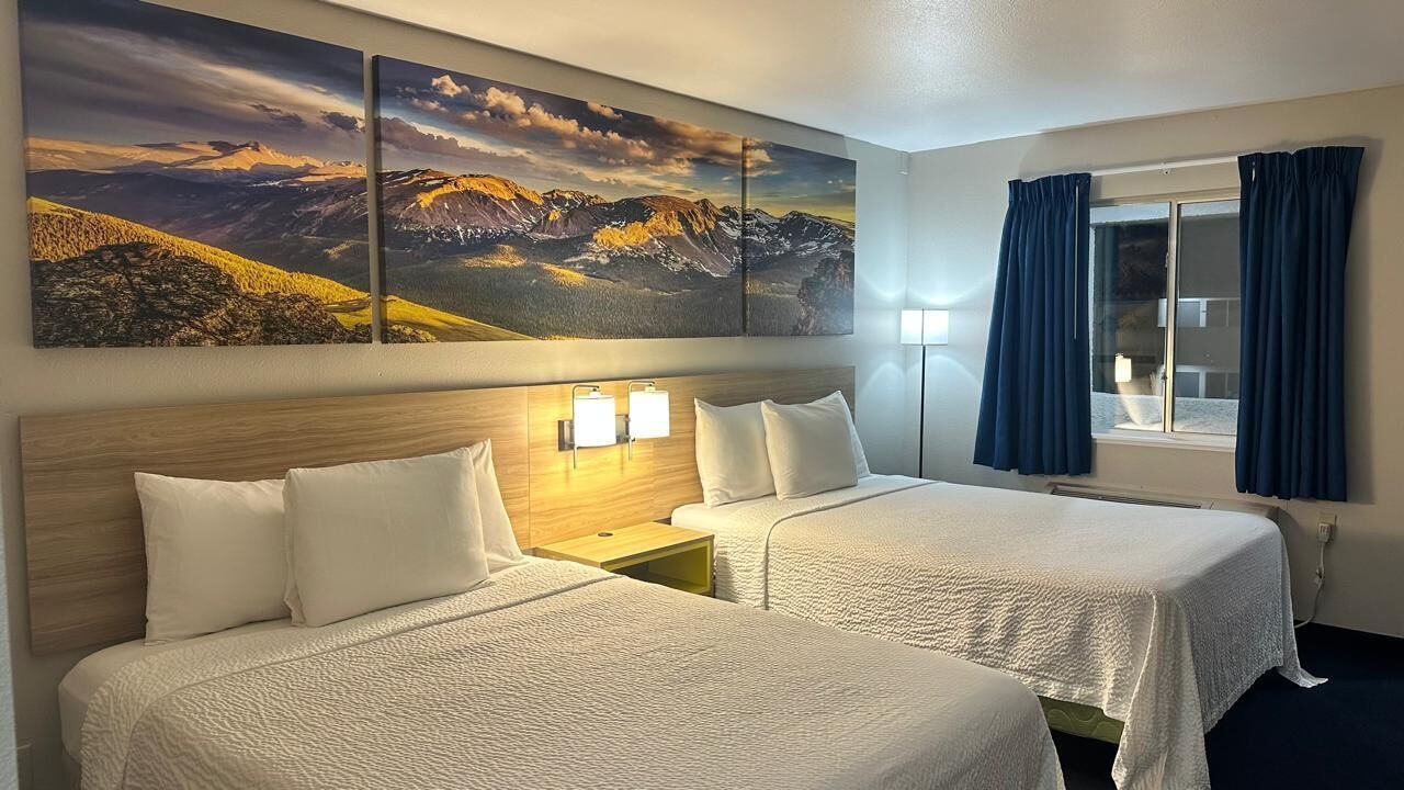 Habitació d'hotel amb dos llits, un panell de capçalera de fusta, una tauleta de nit, làmpades fixades a la paret i un gran quadre paisatgístic sobre els llits.
