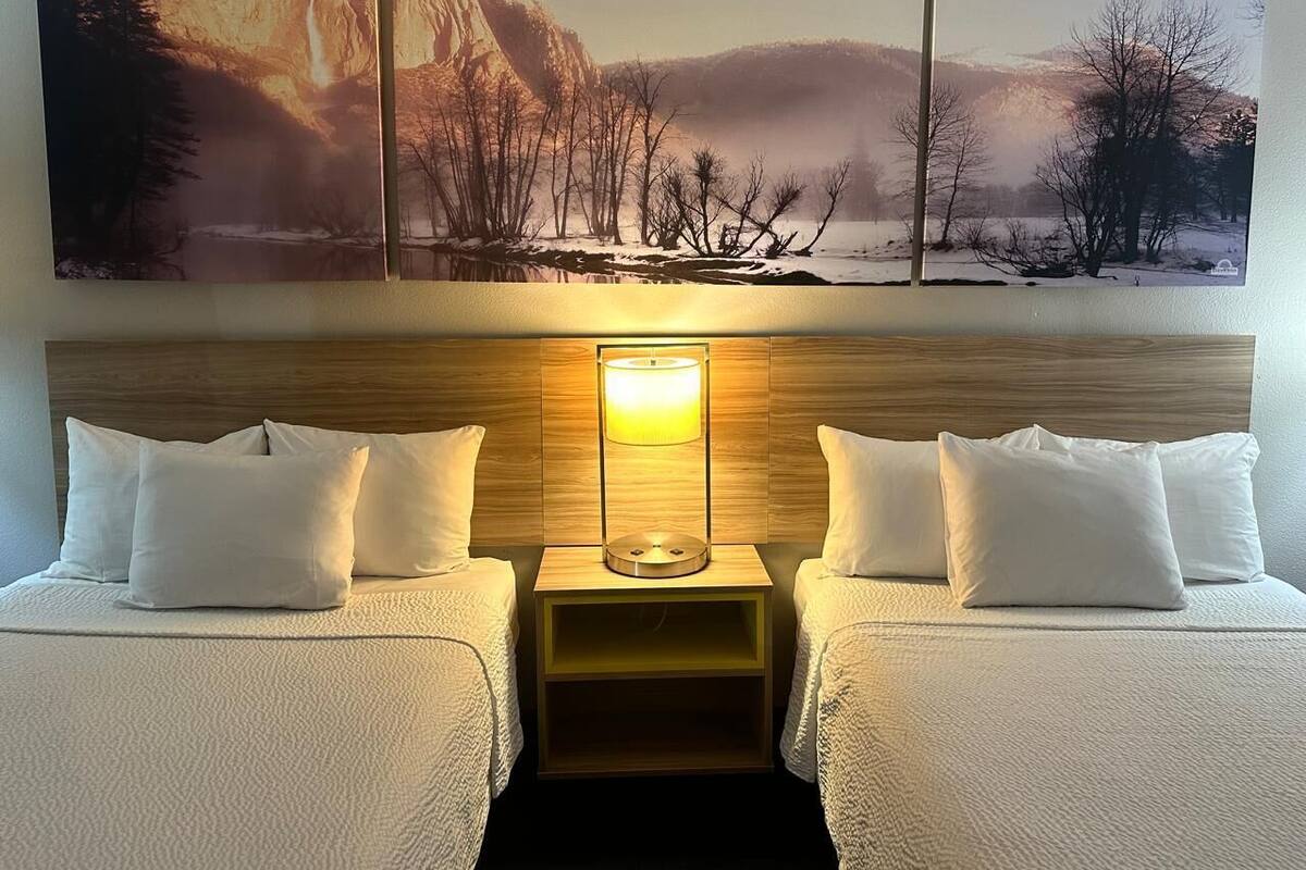 A hotel room with two beds, a wooden headboard, a nightstand with a lamp, and three large landscape paintings above the beds.